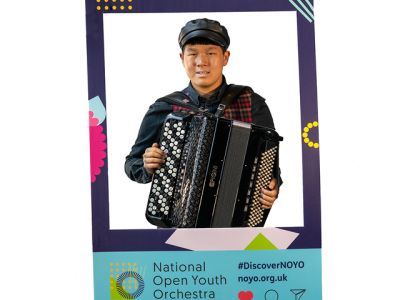 A boy wearing a leather cap, blue shirt and tartan waistcoat holds the accordion. He stands in between a customised National Open Youth Orchestra Instagram frame.